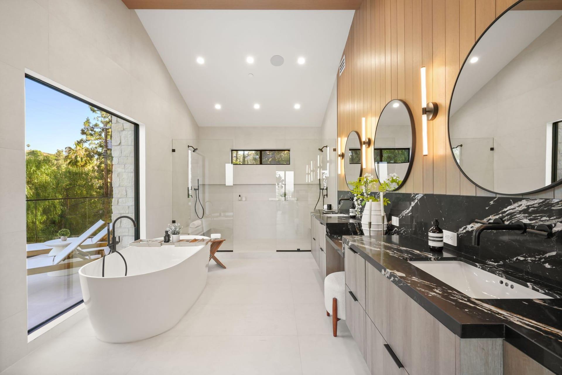 Primary bath with a freestanding tub by a large window, a dark stone double vanity and round mirrors