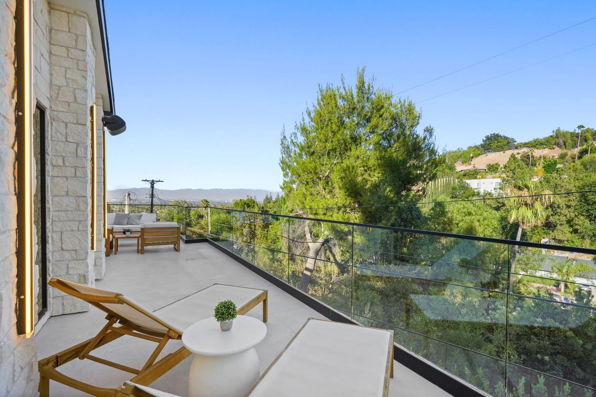 Stone-clad balcony with a glass railing, lounge chairs and a sitting area, looking over wooded hillside to distant mountains