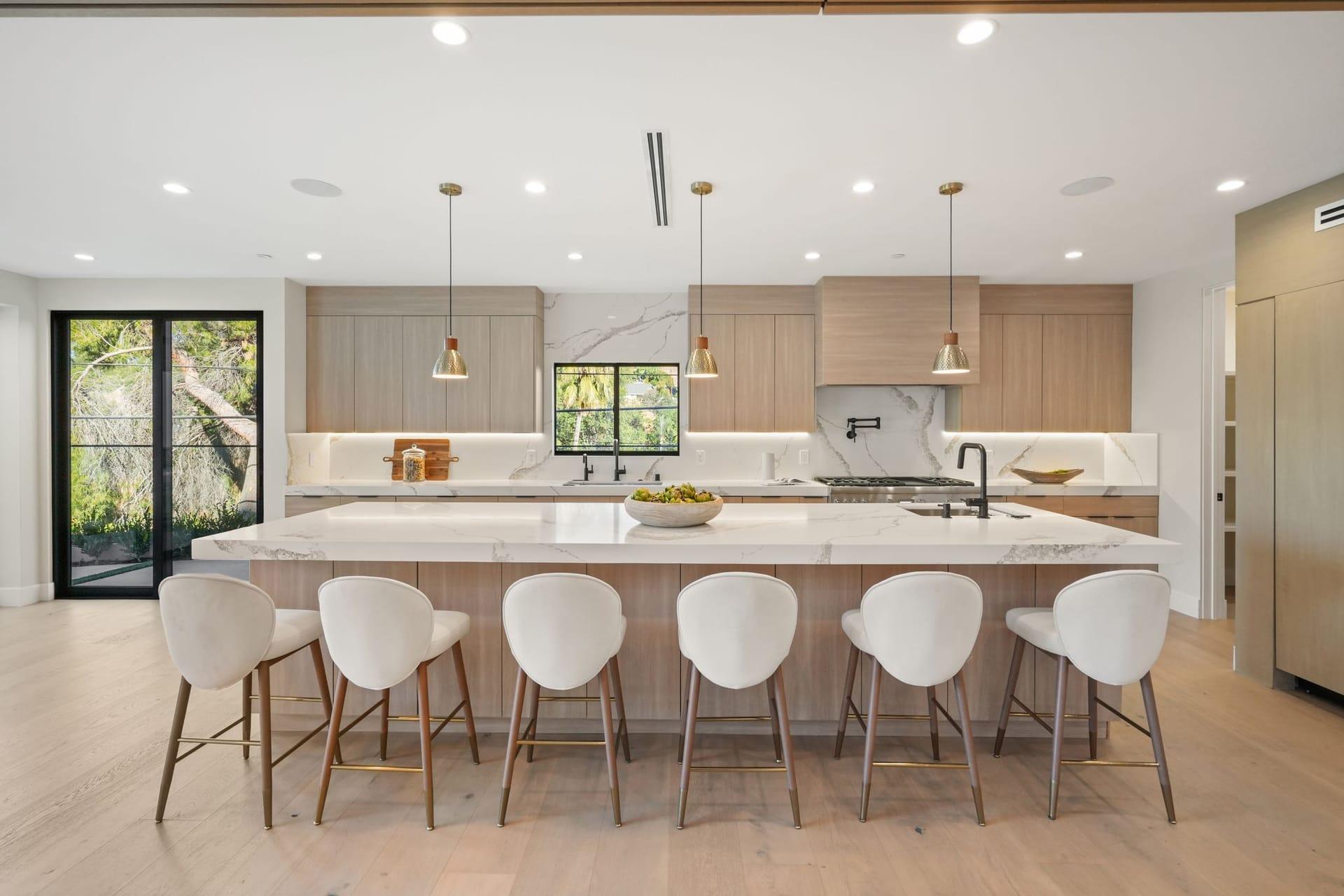 Staged kitchen with a long marble island, counter stools, pendant lights and pale wood cabinetry