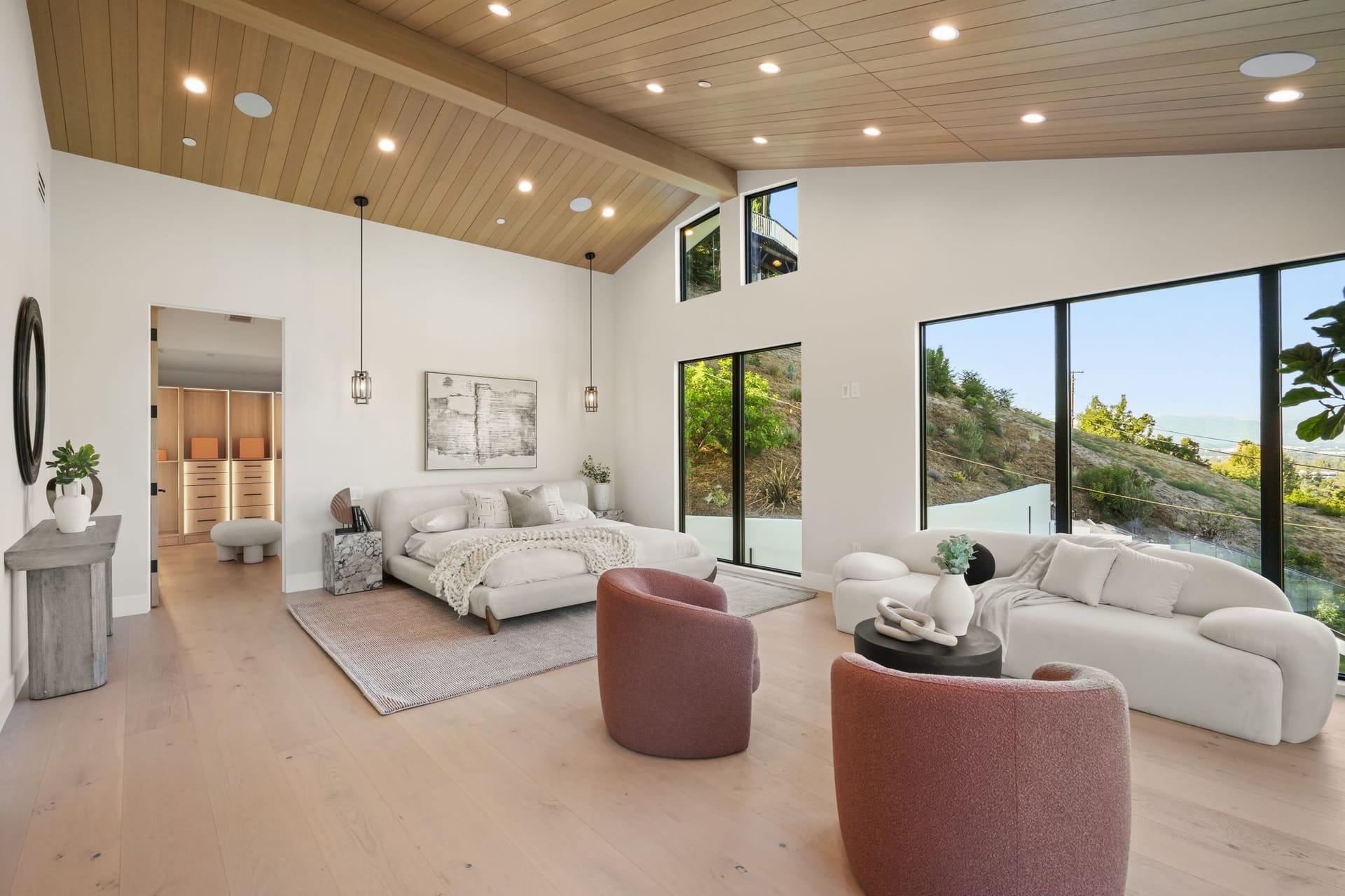 Primary suite under a vaulted timber ceiling, furnished with a bed and a separate sitting area by the windows