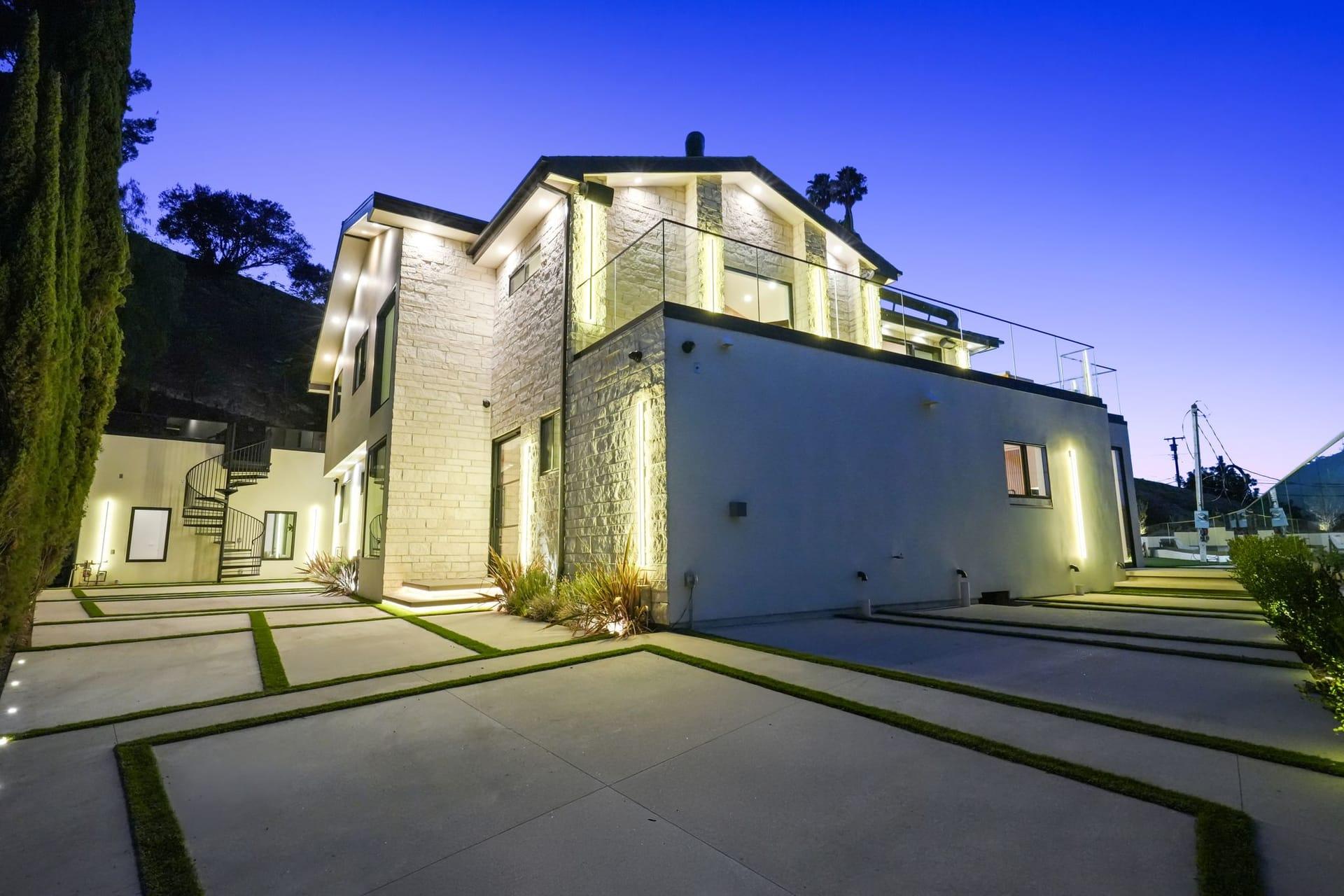Stone-clad side elevation lit at dusk above a paved court set with strips of lawn