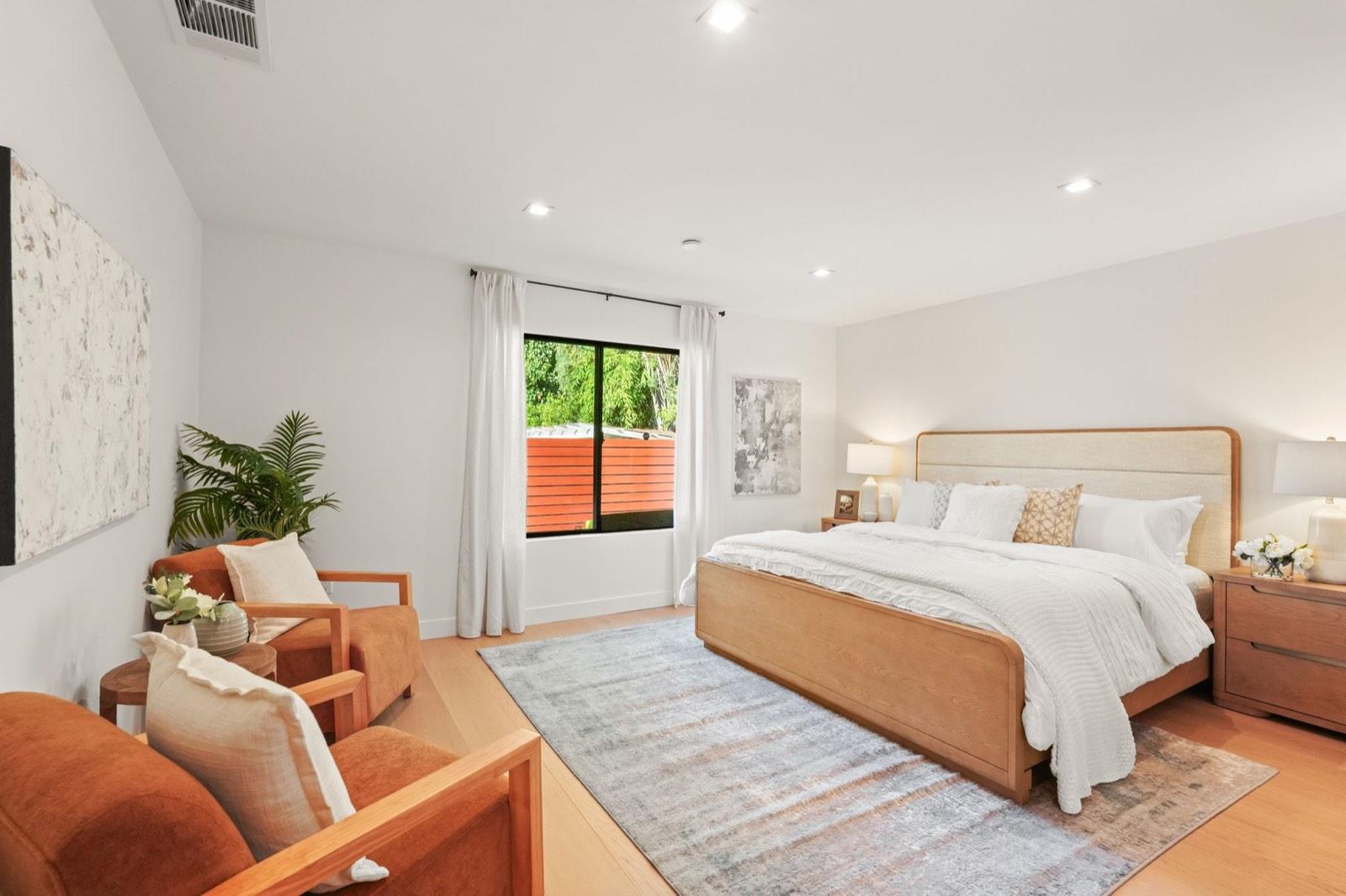 Primary bedroom staged with an upholstered bed, a reading chair and plants by the window