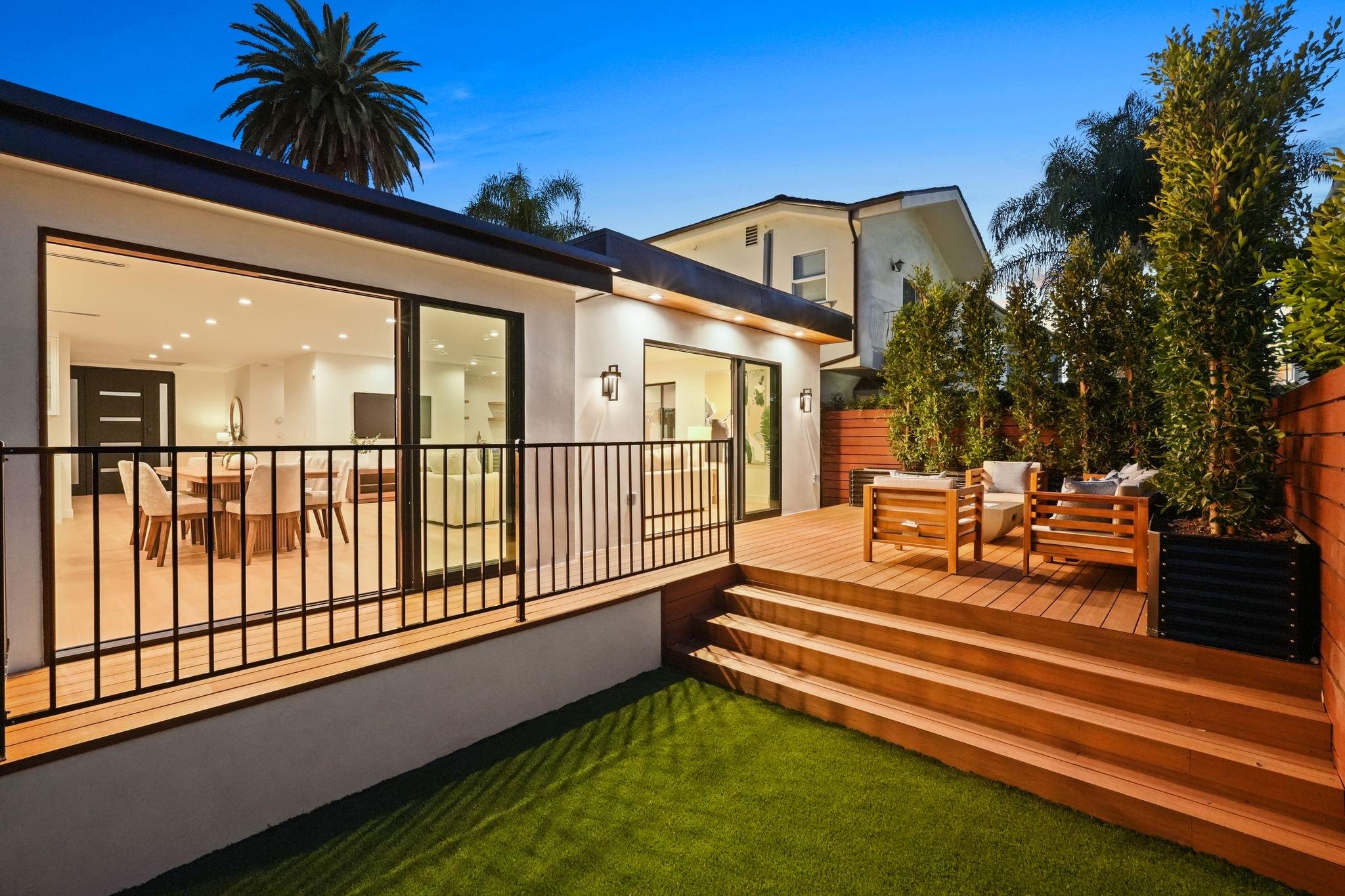 Rear of a remodeled home at dusk, lit through wide glass doors, with a railed timber deck, broad steps and a lawn